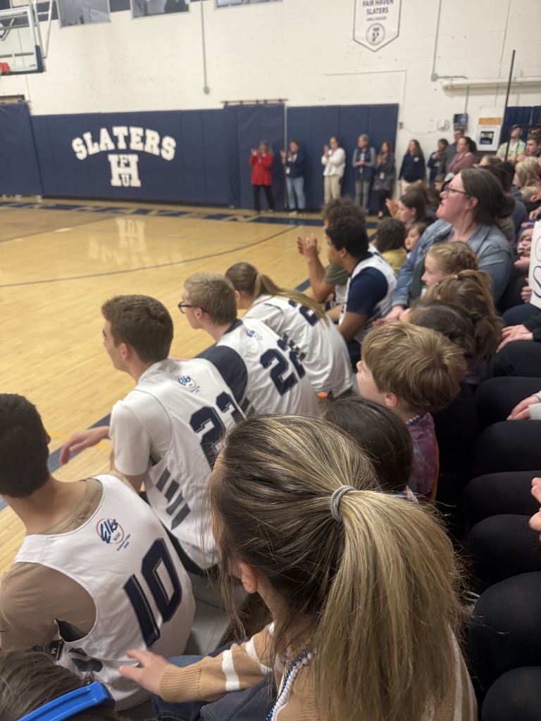 Students and young children sit closely together on gym bleachers during a basketball game. Several players in white jerseys sit in front while younger students wearing beads watch from the seats behind them.