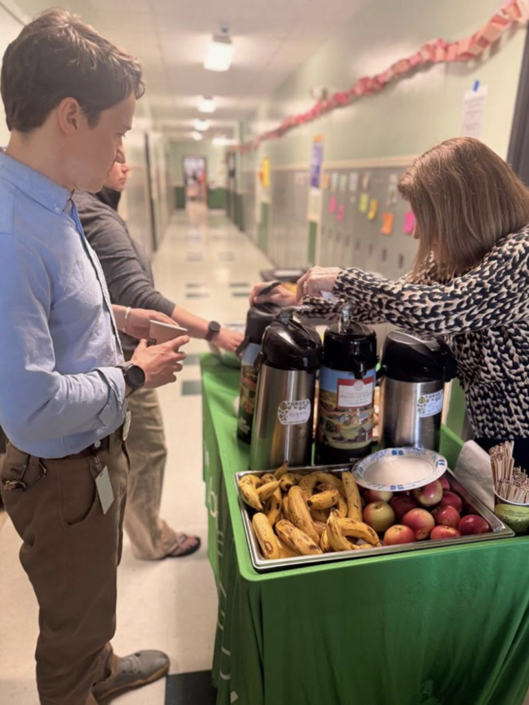 A staff member pours coffee from a thermal dispenser on a green-covered table in a school hallway. Another staff member stands nearby holding a cup. The table is set with bananas, apples, cups, and coffee supplies.
