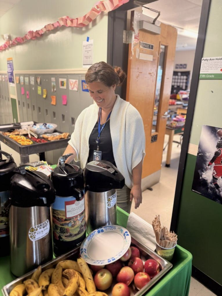 A smiling staff member stands behind a refreshment table with coffee dispensers, fruit, and cups in a school hallway. Classroom doors and student work are visible in the background.