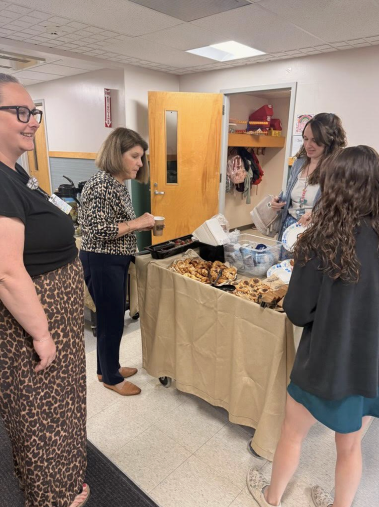 Several staff members gather around a table with pastries and coffee in a school setting. One person pours a drink while others chat and hold plates, enjoying the appreciation treats.