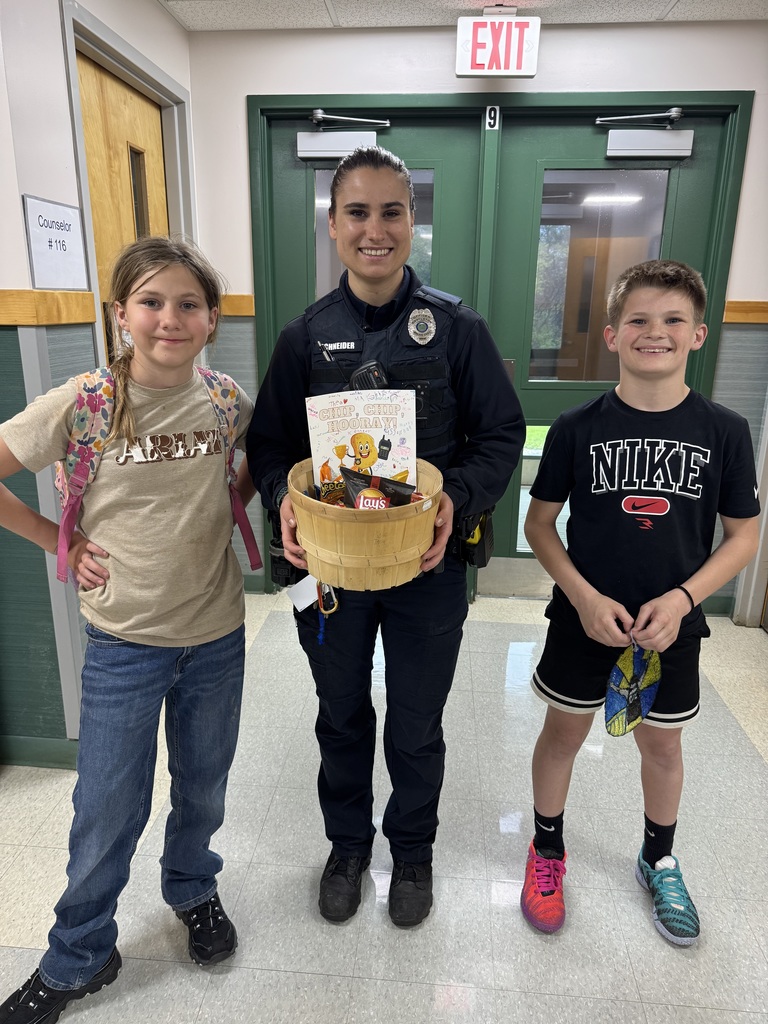 An eye-level shot indoors shows a female police officer smiling, flanked by a female and male student, both smiling, holding a wooden basket of snacks that includes bags of Lays and Cheetos chips, a carton of Goldfish crackers, and a drawing with a smiling potato character that says, “CHIP CHIP HOORAY” above it. The girl is on the left in a khaki shirt that says, “ADRIEN” in block letters and jeans, wearing a multicolored floral backpack. The boy is on the right in a black t-shirt that says, “NIKE” with a logo below and black shorts, holding a yellow and blue mesh mask with both hands, and wearing mismatched sneakers, pink on his right, blue and green on his left. The officer is wearing her police uniform with a radio and holster. behind them is an interior door painted green with double windows, an exit sign above, with the number “9” in a square below the exit sign, and a window to the right of the door. The sign to the left of the door says, “Counselor #116.”