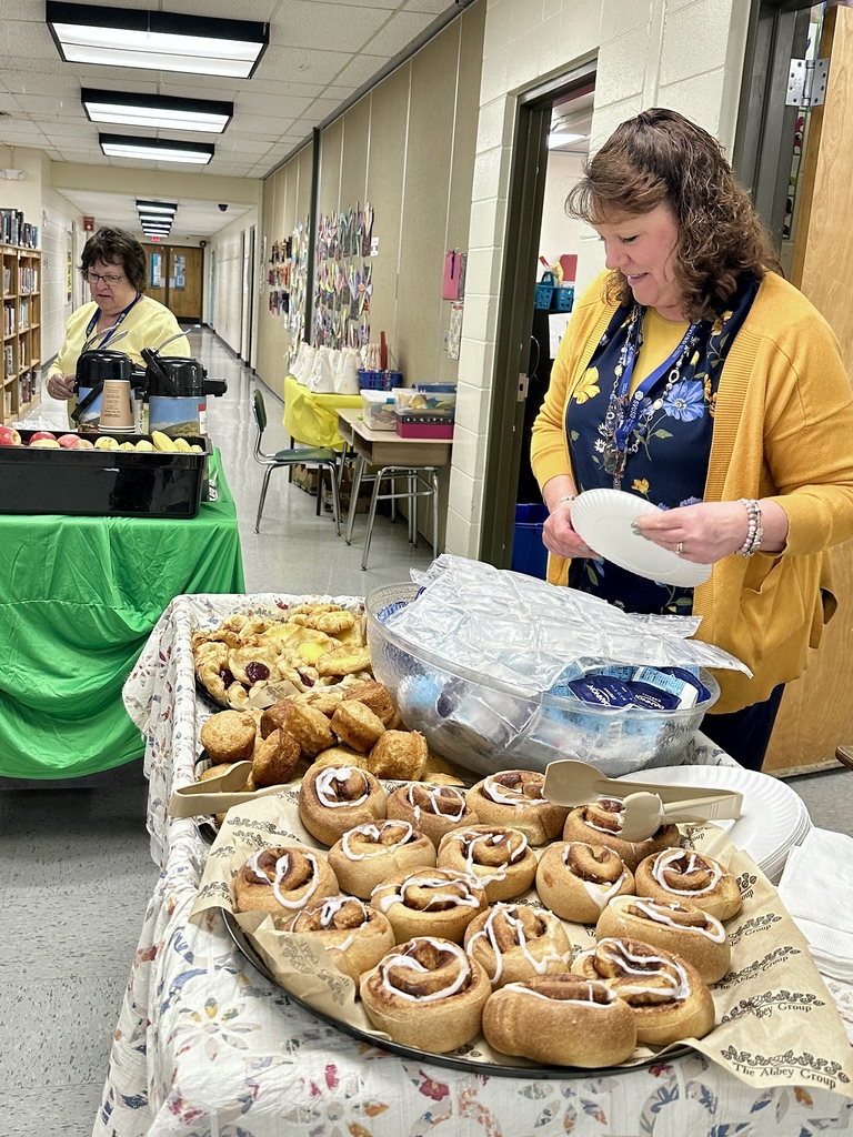 A staff member stands beside a tray of cinnamon rolls and pastries while preparing a plate in a school hallway during Teacher and Staff Appreciation Week.