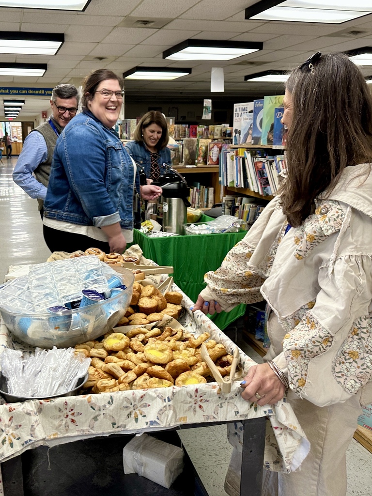 Several staff members smile and talk around a pastry and coffee cart set up in a school library filled with bookshelves.