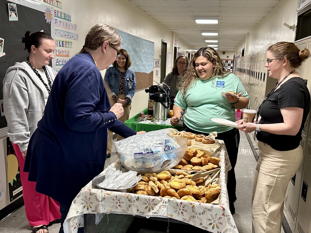 Faculty and staff gather around a breakfast cart filled with pastries, fruit, and coffee in a school hallway while chatting and enjoying refreshments together.