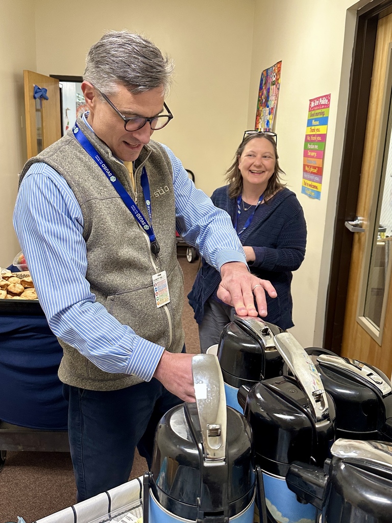 A man pours coffee from a large coffee dispenser while another staff member smiles nearby in a school office area during a faculty appreciation event.
