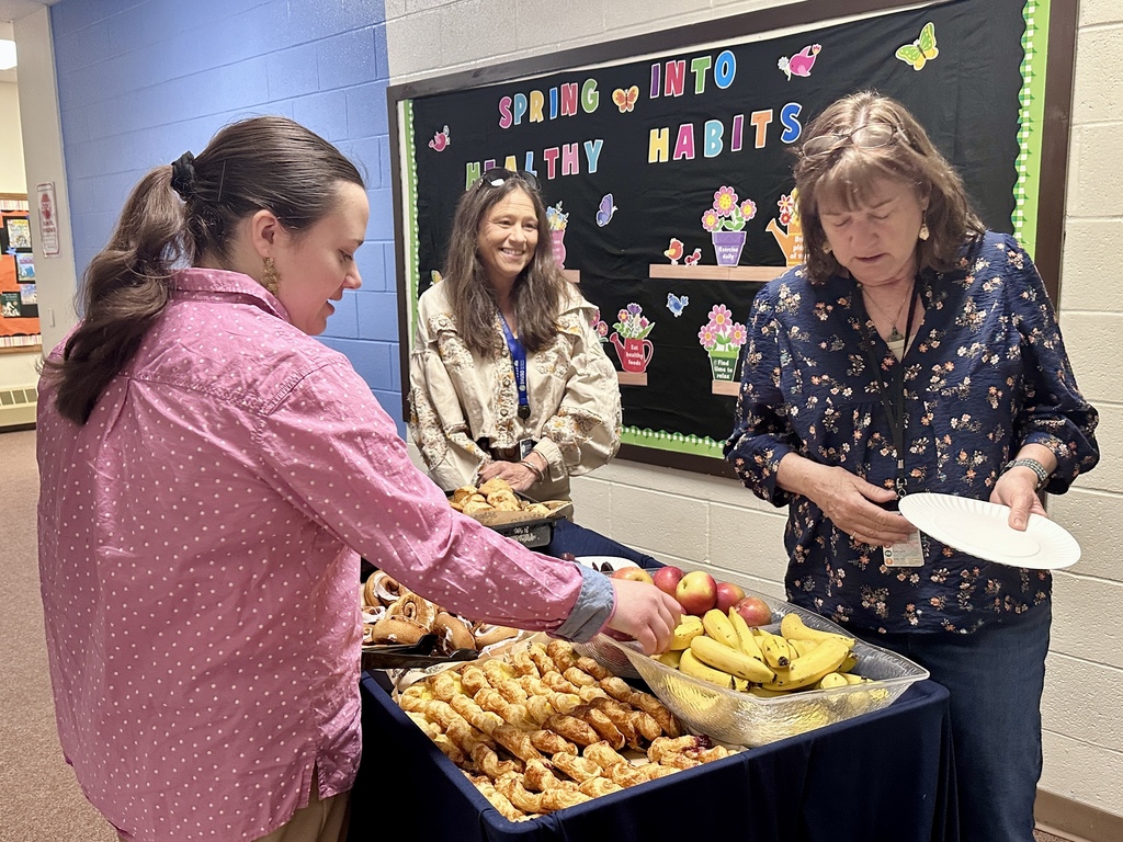 Staff members gather around a pastry cart in a school hallway selecting fruit and breakfast treats. A bulletin board behind them reads “Spring Into Healthy Habits.”