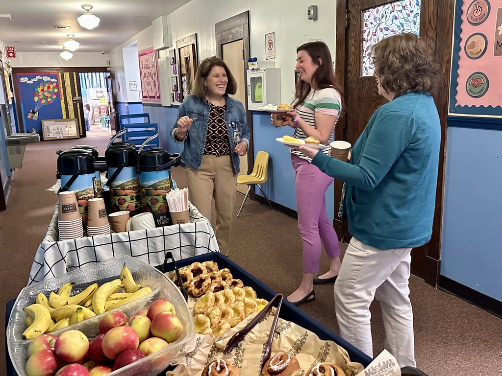 Three staff members stand in a school hallway chatting and smiling near a coffee and pastry cart filled with fruit, pastries, and coffee containers during Teacher and Staff Appreciation Week.