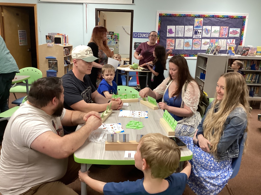Families and children gather around a table in the library playing interactive math games with cards and wooden game boards during a kindergarten family math night.