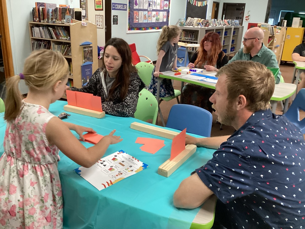 Students and adults sit together at tables in the library playing educational math games with cards and activity boards during a family learning event.