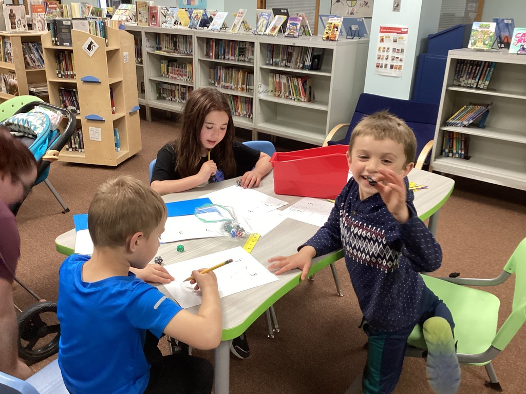 Three young students sit around a table in the library working on number activities with pencils, dice, and paper while one smiling student holds up a game piece.