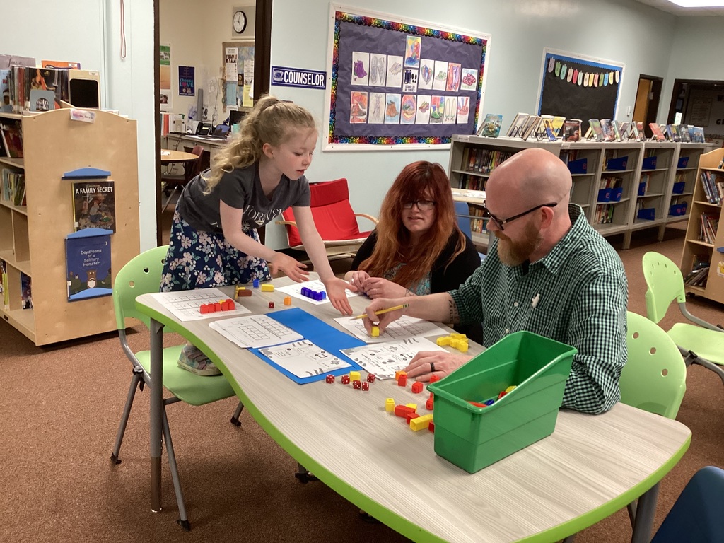 A kindergarten student stands beside two adults at a table in the school library while demonstrating a math game using colorful counting cubes, dice, and worksheets.