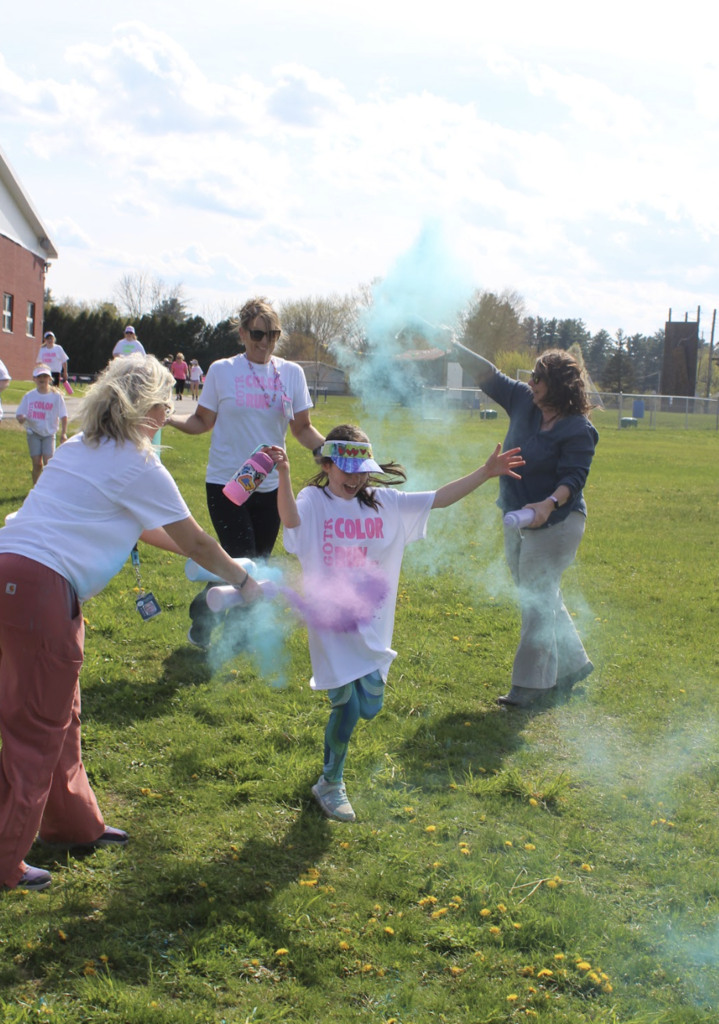 A Girls on the Run participant runs joyfully through blue and purple powder clouds while volunteers toss color from both sides.