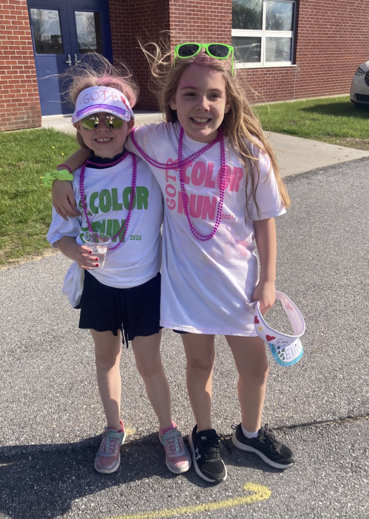 Two young Girls on the Run participants stand side by side outdoors with their arms around each other wearing white Color Run shirts, beads, and sunglasses.