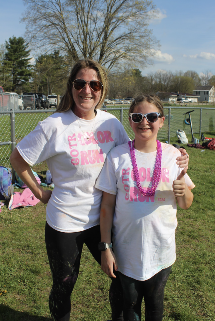 An adult volunteer and a Girls on the Run participant smile together outdoors while wearing matching white Color Run shirts and sunglasses.
