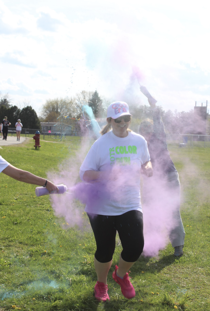 An adult volunteer runs through purple powder during the practice Color Run while smiling and wearing a white Color Run shirt and visor.