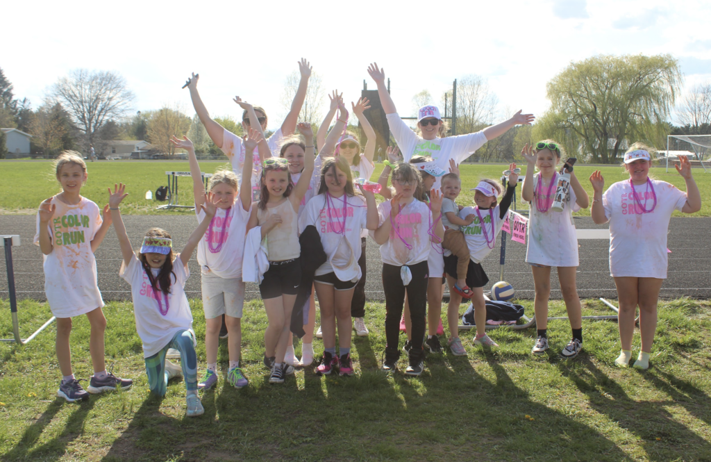 A large group of Girls on the Run participants and volunteers stand together outdoors on a sunny day with their arms raised while wearing white Color Run shirts and colorful accessories.