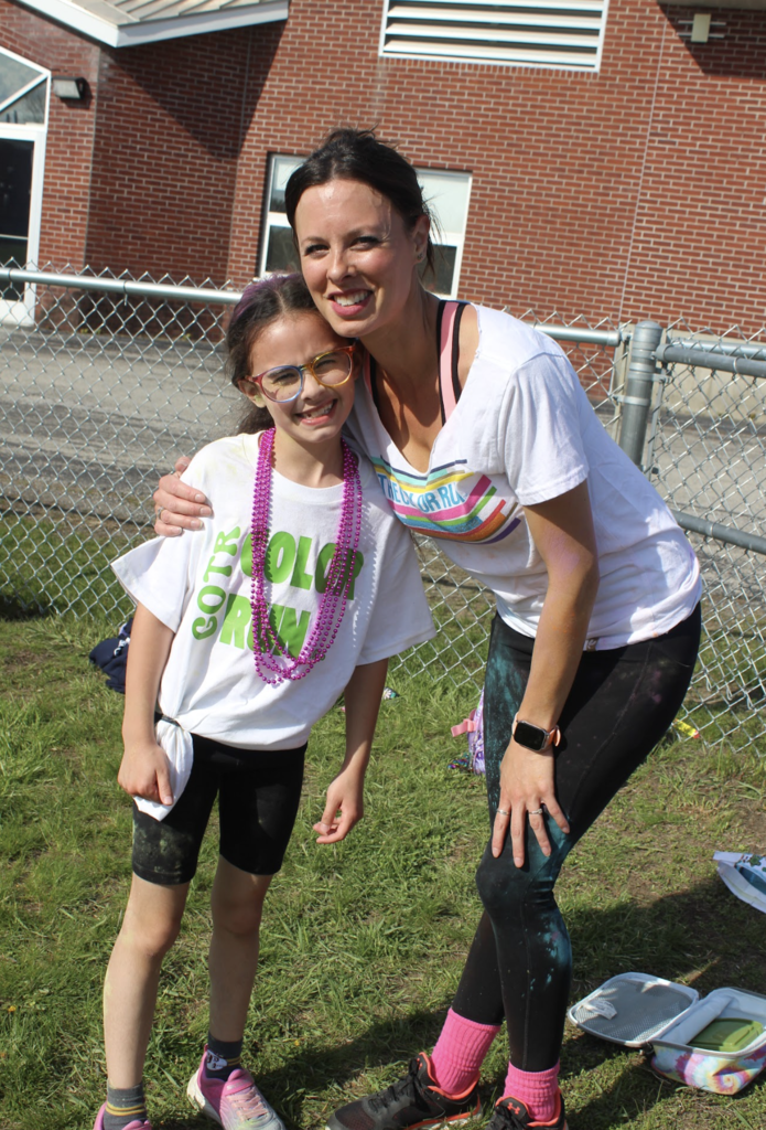 A Girls on the Run participant and an adult volunteer pose together smiling near a fence while wearing white Color Run shirts and colorful beads.