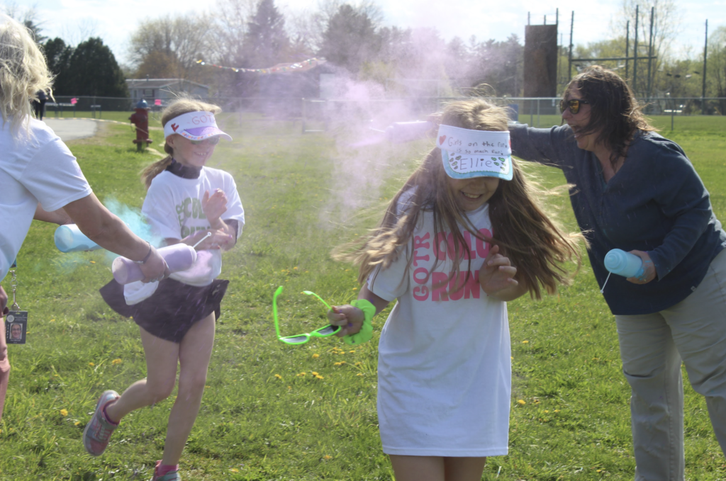 Two Girls on the Run participants run through pink and blue powder clouds while volunteers cheer and toss color during the event.