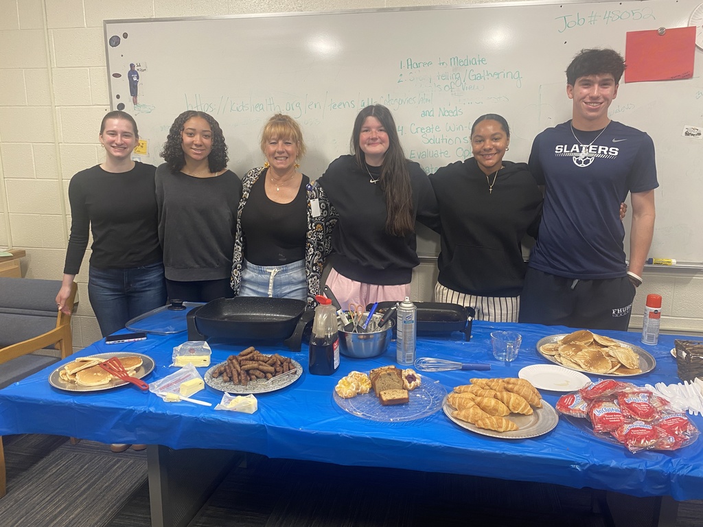 A group of FHUMHS Student Council members and an adult advisor pose behind a table filled with pancakes, sausage, croissants, pastries, and breakfast foods prepared for staff appreciation week.