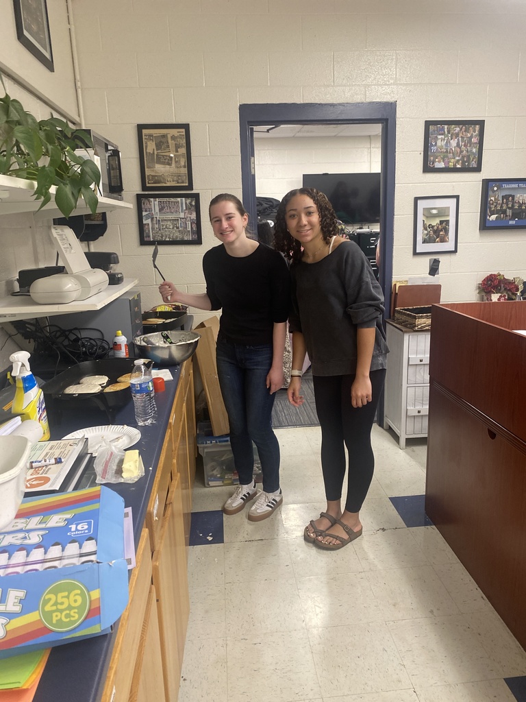 Two FHUMHS students stand smiling near a griddle while cooking pancakes and preparing breakfast items for faculty and staff appreciation.
