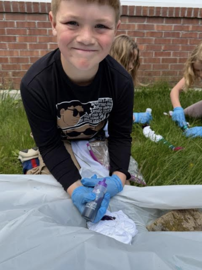 A smiling student wearing blue gloves holds a bottle of dark tie-dye dye while sitting outside near classmates working on tie-dye shirts in the grass.
