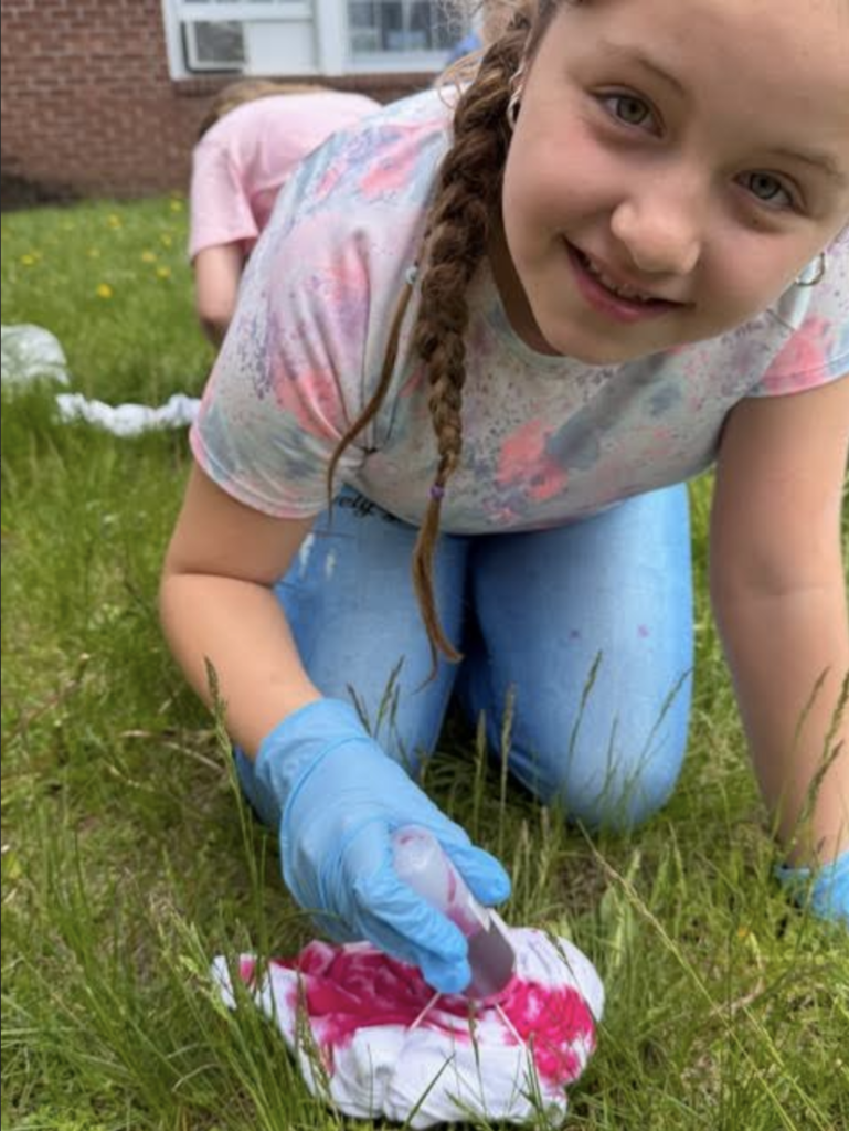 A student wearing blue gloves leans close to the camera while carefully applying bright pink dye to a folded white shirt during a tie-dye activity outside.