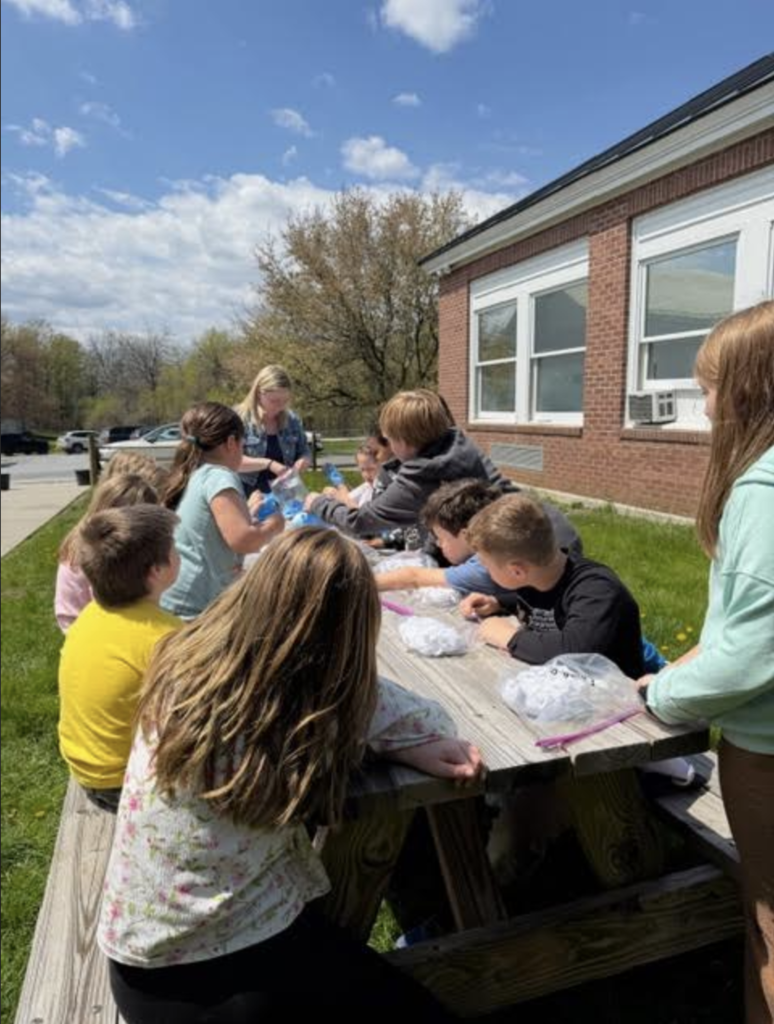 A group of students and an adult gather around a picnic table outside the school while working together on tie-dye shirt projects on a sunny day.
