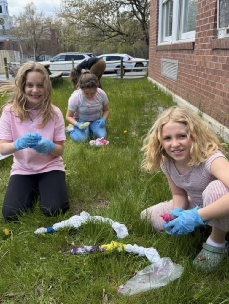 Three students kneel in the grass outdoors while using colorful dyes to create tie-dye shirts during an outdoor activity beside a brick school building.