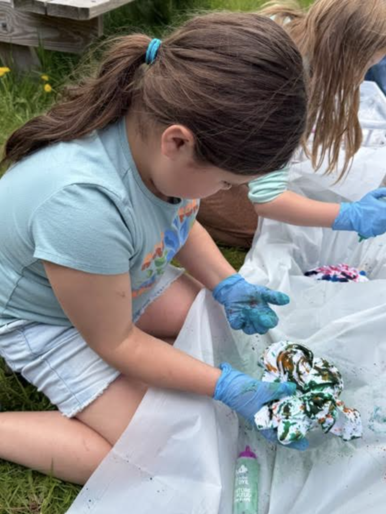 A student wearing blue gloves sits on the grass outdoors while creating a tie-dye shirt with green and orange dye. Another student works on a project beside them.