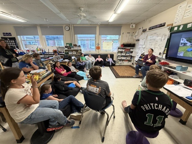 A guest speaker stands at the front of an elementary classroom speaking to students seated on the floor and at desks while a digital screen displays presentation materials.