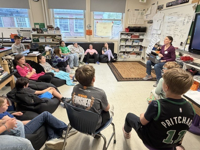 A guest speaker stands at the front of an elementary classroom speaking to students seated on the floor and at desks while a digital screen displays presentation materials.