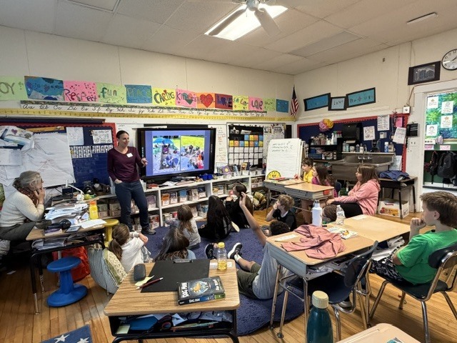 A wide view of a classroom shows students gathered around a guest speaker presenting information on a large digital screen during an interactive lesson.