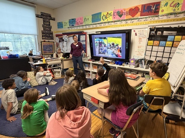 A wide view of a classroom shows students gathered around a guest speaker presenting information on a large digital screen during an interactive lesson.