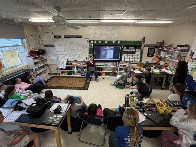 A wide view of a classroom shows students gathered around a guest speaker presenting information on a large digital screen during an interactive lesson.