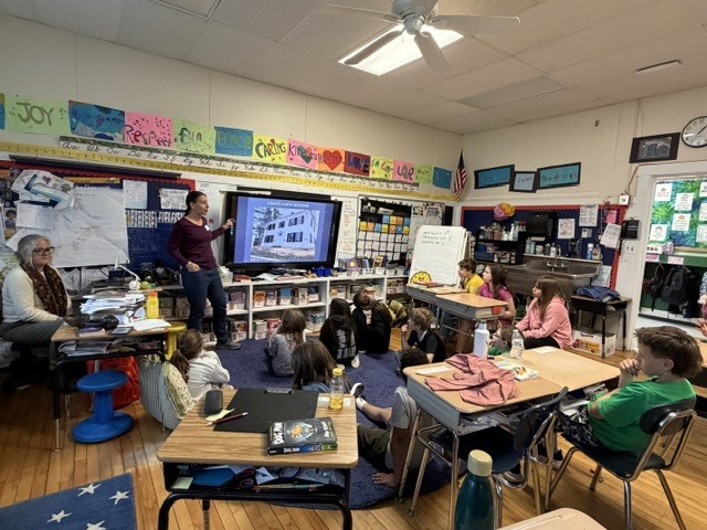 A wide view of a classroom shows students gathered around a guest speaker presenting information on a large digital screen during an interactive lesson.