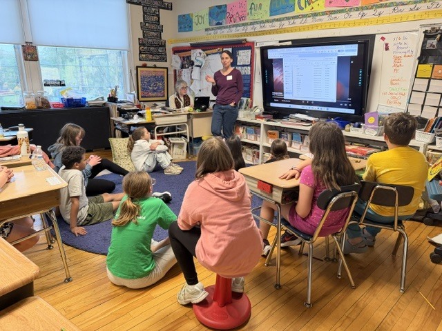 Middle school students sit around a classroom listening to a guest speaker seated near a large interactive screen during a presentation and discussion activity.