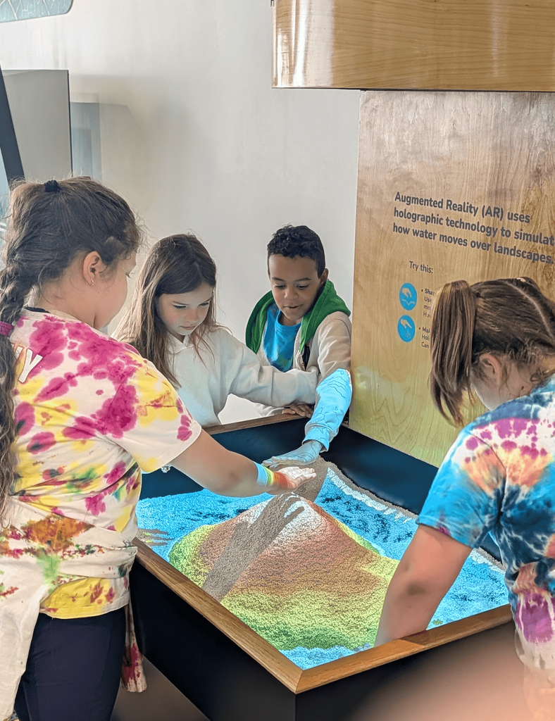 Students gathered around an augmented reality sand table use their hands to shape mountains and valleys while projected colors simulate water and landscapes.