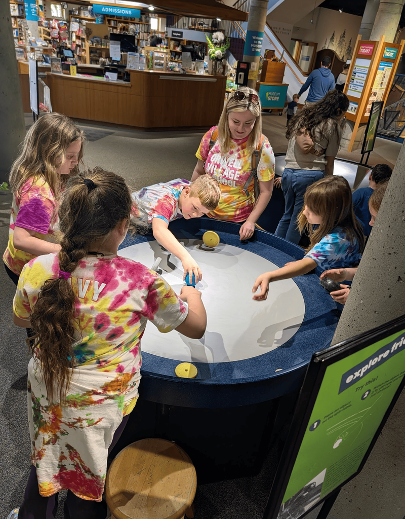 Students and an adult chaperone gather around a circular interactive exhibit at the museum while experimenting with spinning objects and motion activities.