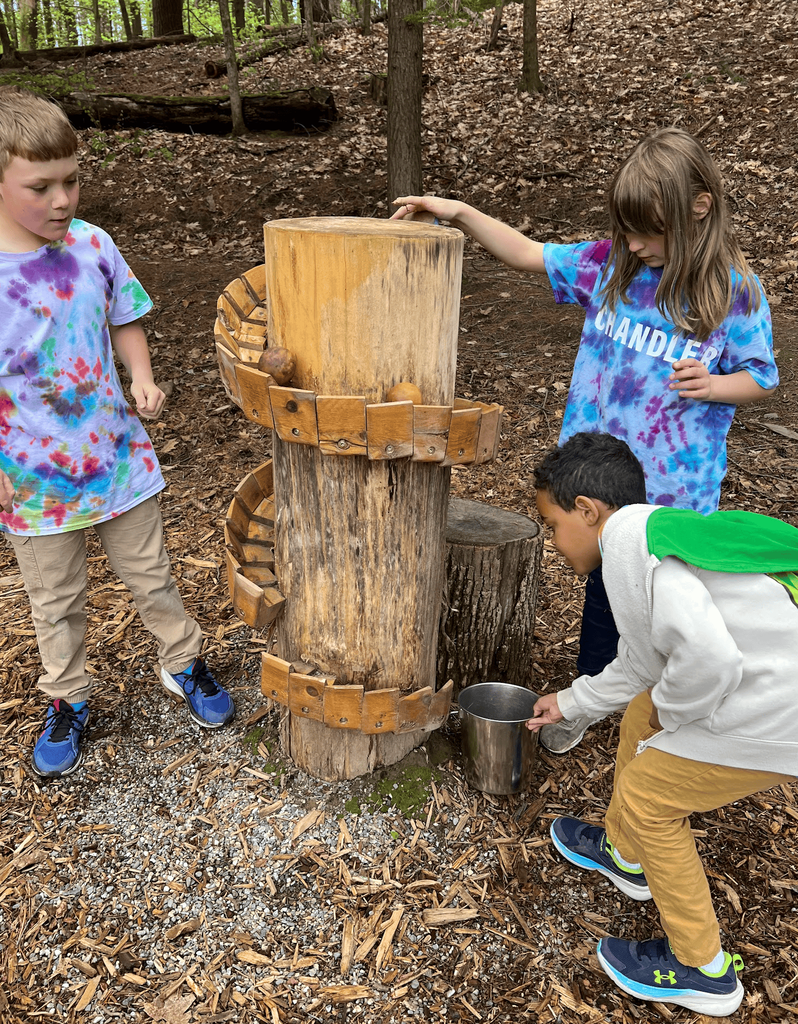 Three students explore a wooden gravity and water-themed outdoor exhibit by guiding objects through spiral tracks in a wooded area.