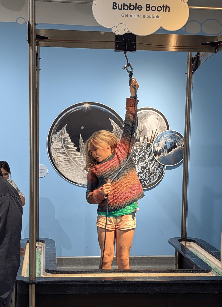 A student stands inside the Montshire Museum bubble booth exhibit while pulling a rope mechanism to create a giant bubble around herself.