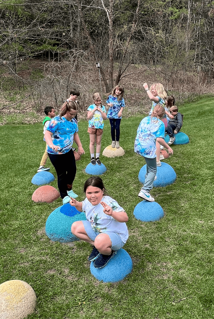 Third grade students wearing tie-dyed shirts balance and play on colorful dome-shaped playground structures outdoors.