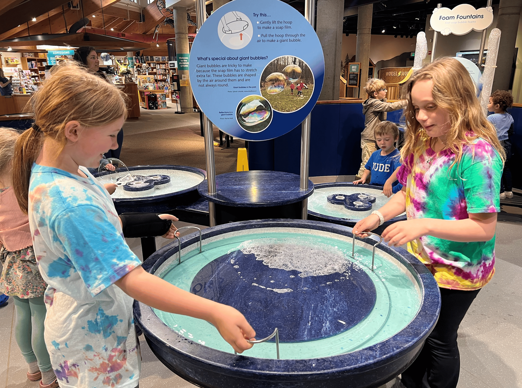 Two students wearing colorful tie-dyed shirts use giant bubble-making stations at the Montshire Museum while other children explore exhibits in the background.