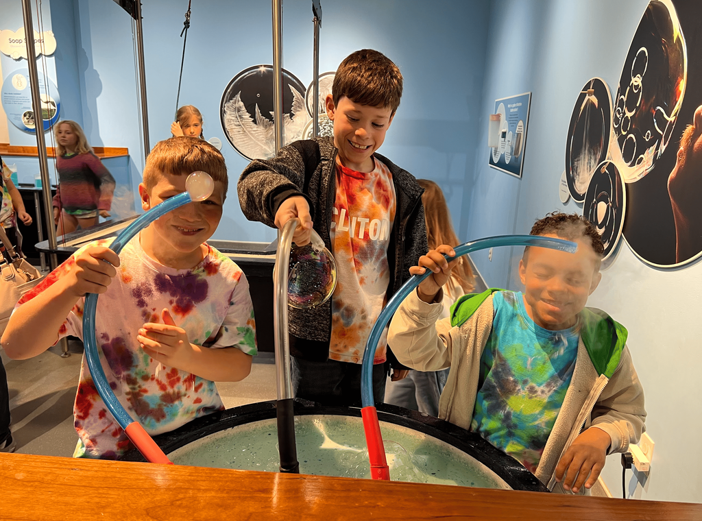 Three smiling students wearing matching tie-dyed shirts explore bubble-making activities together at a hands-on science exhibit.