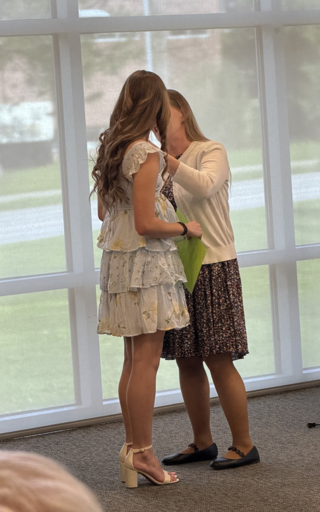 A mentor teacher pins a student teacher during the pinning ceremony while the student teacher stands in a light blue floral dress holding papers.