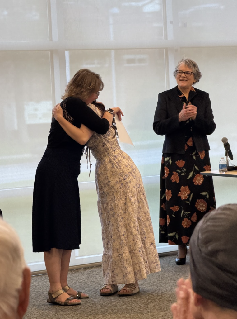 A mentor teacher hugs a student teacher during the pinning ceremony while another woman stands nearby clapping.