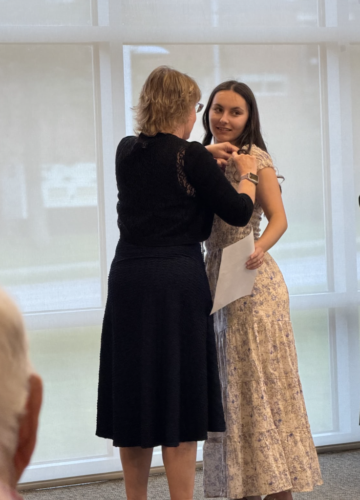A mentor teacher pins a student teacher wearing a long floral dress during the ceremony as audience members watch.