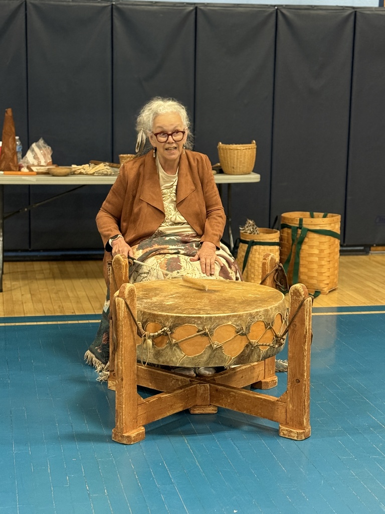Miss Lucy Neel sits behind a large traditional drum in the gym while demonstrating drumming techniques and sharing Abenaki traditions with students.