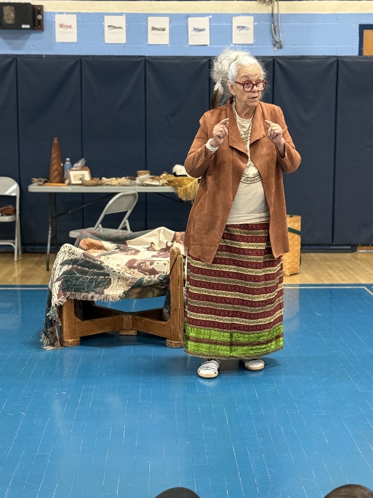 Miss Lucy Neel stands in the gym speaking to students about Abenaki history and traditions while cultural items are displayed behind her.