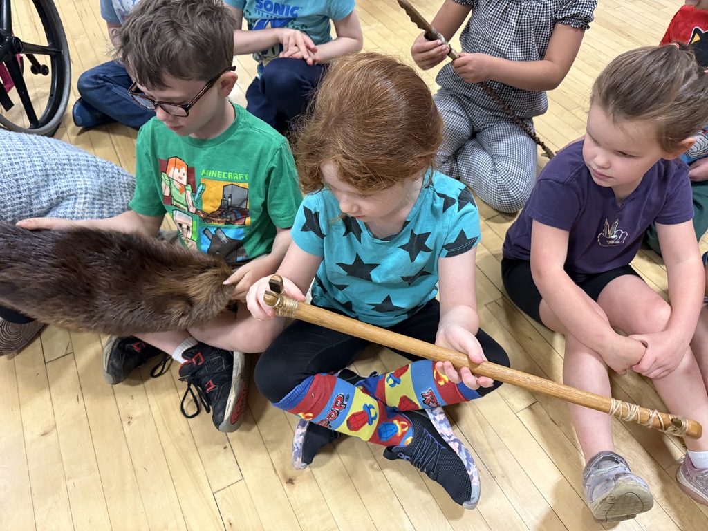Students sit on the gym floor examining traditional cultural items, including a fur and handcrafted tools, during an Abenaki culture presentation.