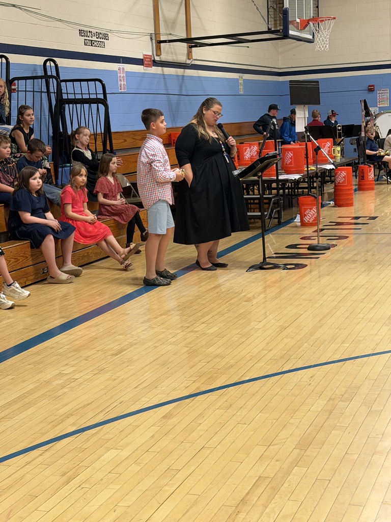 Mrs. Hutchins stands beside a student at a microphone during an awards or recognition portion of the FHGS spring concert while students sit nearby on the bleachers.